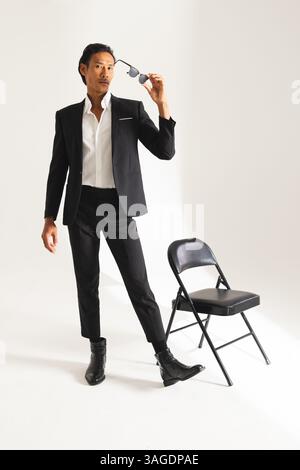 Selbstbewusster Mann in stilvollem Anzug, der eine Sonnenbrille hält und neben dem Stuhl steht. Mode, Business, Selbstvertrauen, formell, trendy, Stil Stockfoto