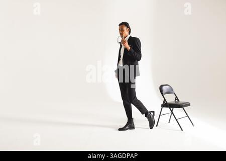 Stilvoller Mann im Anzug, der eine Brille hält und selbstbewusst in der Nähe des Stuhls im Studio posiert. Mode, Eleganz, Modellierung, Raffinesse, Kleidung, Vertrauen Stockfoto