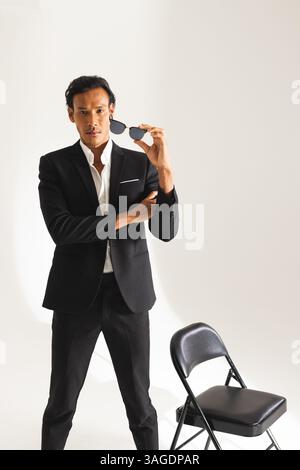 Selbstbewusster Mann im Anzug, der eine Sonnenbrille hält und neben einem schwarzen Stuhl im Studio steht. Business, Profi, Mode, Eleganz, Selbstvertrauen, Hochformat Stockfoto