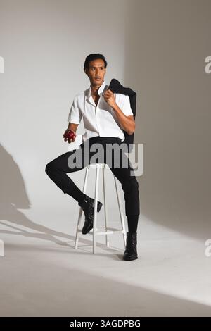 Selbstbewusster Mann in stilvollem Outfit, der Apfel hält und im Studio auf dem Hocker sitzt. Mode, Selbstvertrauen, Lifestyle, Bekleidung, Porträt, männlich Stockfoto
