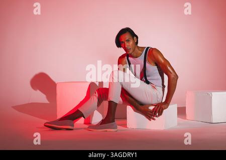 Selbstbewusster Mann in stilvollem Outfit, der im Studio auf weißen Würfeln posiert. Mode, Selbstvertrauen, trendy, Bekleidung, modern, städtisch Stockfoto