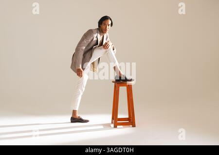 Selbstbewusster Mann in stilvollem Outfit, der im Studio mit dem Fuß auf dem Hocker posiert. Mode, Stil, Selbstvertrauen, Herrenmode, Porträt, trendig Stockfoto
