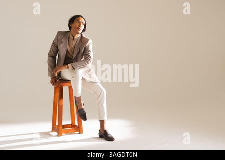 Stilvoller Mann in elegantem Outfit, der auf dem Hocker sitzt und nachdenklich absieht. Mode, Stil, Raffinesse, Herrenmode, trendy, nachdenklich Stockfoto
