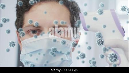 Bild von covid-19-Zellen über dem Menschen in Gesichtsmaske mit Temperaturkontrolle Stockfoto