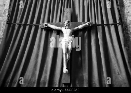 Altes hölzernes Kruzifix von Jesus Christus isoliert auf Vorhängen in der mittelalterlichen Kirche. Schwarzweißes historisches Foto. Stockfoto