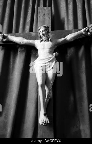 Altes hölzernes Kruzifix von Jesus Christus isoliert auf Vorhängen in der mittelalterlichen Kirche. Schwarzweißes historisches Foto. Stockfoto