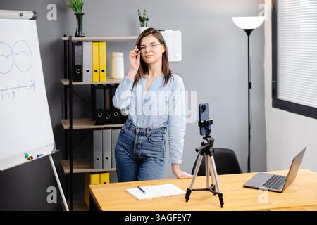 Junge Frau Mentorin nimmt Präsentationen auf, Webinar über Smartphone auf Stativ stehend, Lehrer Coach erklärt, hält Online-Unterricht, mit Telefon-Webc Stockfoto