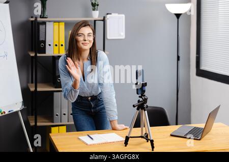 Positive Bloggerin, die auf einem Stativ an der Smartphone-Webcam spricht, einen Beitrag für den Vlog aufnimmt. Online-Lehrer, Coach, Webinar zu Hause. Pressen Stockfoto