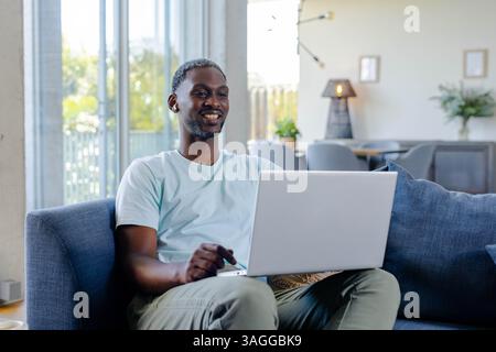 Lächelnder Afroamerikaner, der zu Hause einen Laptop auf der Couch benutzt und die Freizeit genießt. Technologie, Fernarbeit, Entspannung, Lifestyle, Konnektivität, CA Stockfoto