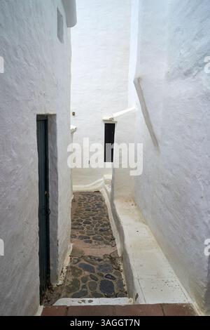 Eine schmale, weiß getünchte Gasse in Binibeca, Menorca, mit Kopfsteinpflaster und dunklen Türen, die eine ruhige und malerische Aussicht bieten. Stockfoto