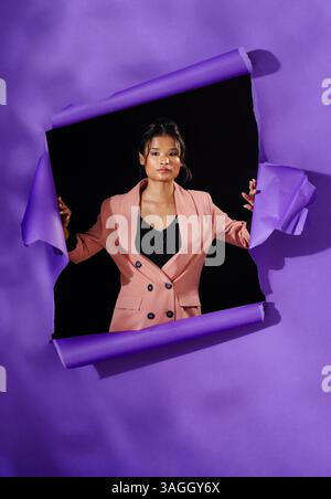 Selbstbewusste asiatische Frau in stilvollem Blazer, die durch gerissenes lila Papier auftaucht. Mode, Empowerment, Selbstvertrauen, mutig, Outfit, trendig Stockfoto