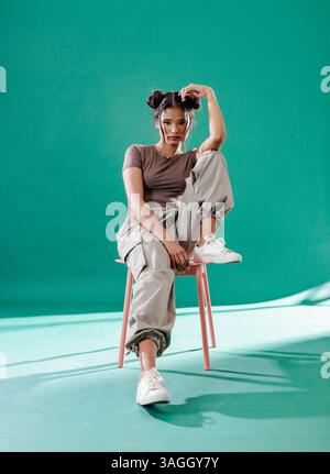 Junge Frau im lässigen Outfit, die selbstbewusst auf dem Stuhl im Studio sitzt. Mode, Selbstvertrauen, Porträt, modern, Lifestyle, Einstellung Stockfoto