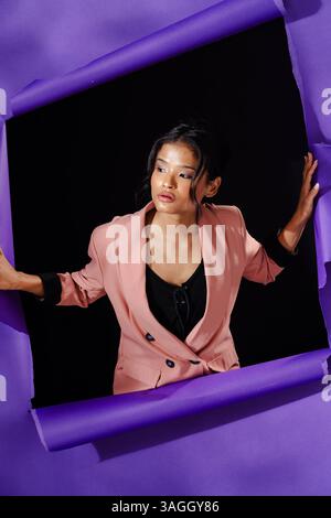 Frau in stilvollem rosa Blazer, die selbstbewusst vor gerissenem lila Papierhintergrund posiert. Mode, Stil, trendig, lebendig, modern Stockfoto