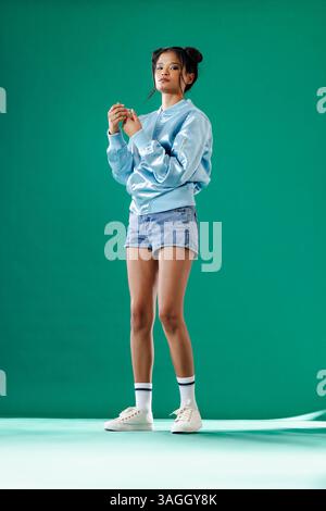 Junge Frau in blauer Jacke und Jeansshorts posiert selbstbewusst im Studio. Mode, Style, Selbstvertrauen, Bekleidung, Fotoshooting, trendig Stockfoto
