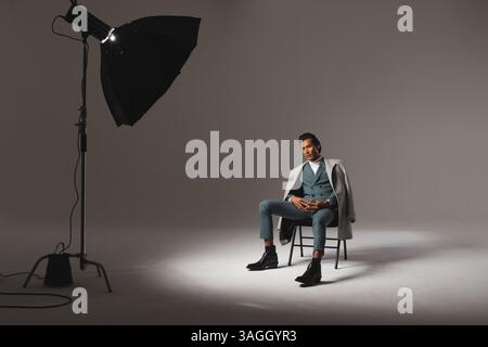 Selbstbewusster Mann in stilvollem Anzug, der auf einem Stuhl unter Studiobeleuchtung sitzt. Mode, Selbstvertrauen, Eleganz, Herrenbekleidung, Geschäftsmann, Hochformat Stockfoto