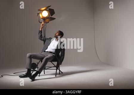 Stilvoller Mann im Anzug, der das Studio-Licht anpasst, während er auf dem Stuhl sitzt. Fotografie, Beleuchtung, Porträt, professionell, Mode, Kreativität Stockfoto