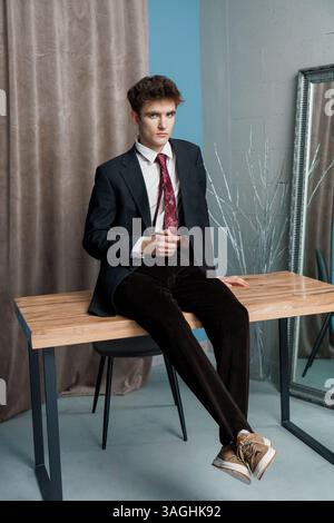 Junger, stilvoller Mann in schwarzem Anzug, gemusterter Krawatte und Samthose, der lässig auf dem Tisch sitzt und drinnen die Beine überkreuzt. Stockfoto