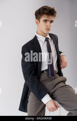Junger Mann in formeller Kleidung posiert selbstbewusst, trägt eine dunkle Anzugsjacke, ein weißes Hemd und eine gemusterte lila Krawatte vor einem neutralen Hintergrund. Stockfoto