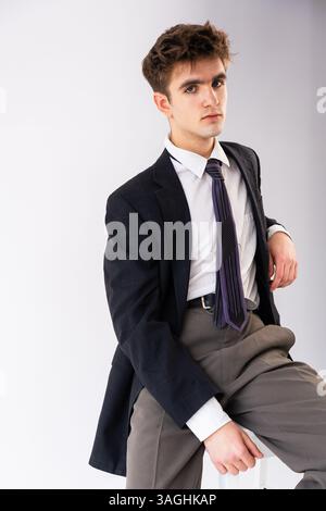 Junger Mann in formeller Kleidung, der sich drinnen vor einem hellgrauen Hintergrund posiert, mit einer dunklen karierten Anzugsjacke, einem weißen Hemd und einer lila Krawatte. Stockfoto