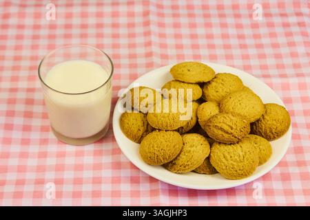 Ein Teller mit Keksen sitzt auf einer rosa-weiß karierten Tischdecke neben einem Glas Milch. Die Kekse sind pyramidenförmig angeordnet, mit der oberen Kappe Stockfoto