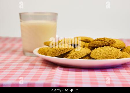 Ein Teller mit Keksen sitzt auf einer rosa-weiß karierten Tischdecke neben einem Glas Milch. Die Kekse sind pyramidenförmig angeordnet, mit der oberen Kappe Stockfoto