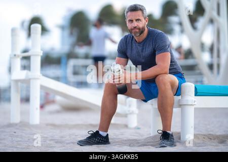 Sport-Fitness-Mann, der draußen auf einer Bank ruht. Reifes Fitnesssportmodell, das nach dem Training auf einer Bank sitzt. Ein Sportmann macht nach dem Fitnesstraining eine Pause Stockfoto