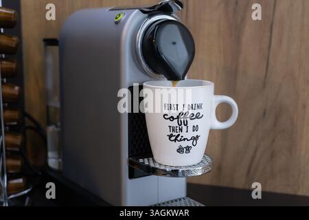 Nahaufnahme einer Krups Nespresso-Kaffeemaschine, die Espresso in eine Tasse gießt, mit dem Text zuerst trinke ich Kaffee, dann mache ich Dinge. Stockfoto