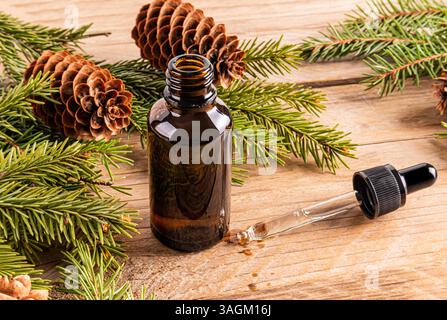 Eine offene dunkle Glasflasche und eine mit Fichtenöl gefüllte Pipette auf einem hölzernen Hintergrund zwischen Fichtenzweigen und -Kegeln. Aromatherapie, Spa-Anwendungen Stockfoto