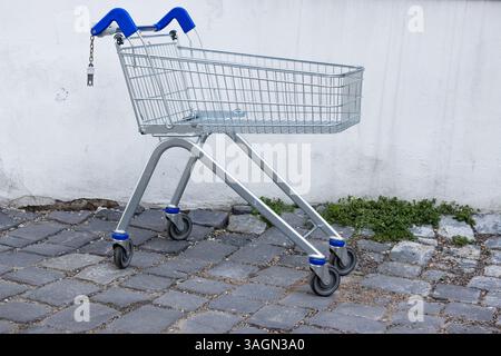Verlassener leerer Einkaufswagen aus Metall mit blauem Griff, der im Hinterhof des Ladens an der schmutzigen Betonwand an der Kopfsteinpflasterstraße stand. Alte dreckige Grunge wa Stockfoto