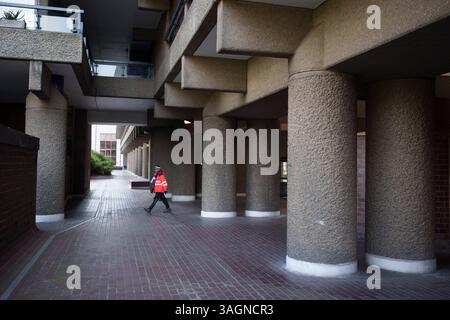 Ein Postbote der Royal Mail liefert am 8. April 2025 in London, England, unter der Architektur der Betonsäulen im brutalistischen Stil des Barbican Estate Development in der City of London. Als Ersatz für den schwer bombardierten Teil der Stadt Cripplegate während des Blitz im 2. Weltkrieg begann der Bau des Barbican Estate 1965 und dauerte 11 Jahre. Der Komplex wurde von den Architekten Chamberlin, Powell und Bon entworfen und heute beherbergt das 40 Hektar große Anwesen mehr als 4.000 Einwohner, die in über 2.014 Wohnungen leben. Stockfoto