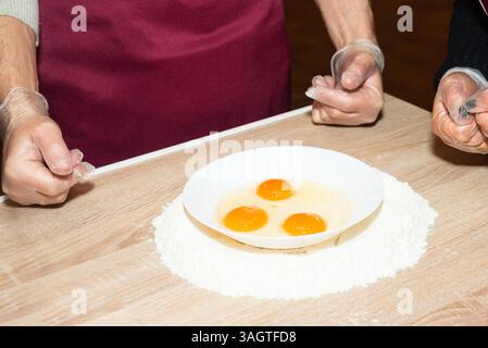Drei rohe Eier in einer weißen Schüssel, die auf einem Hügel Mehl auf einer Holzoberfläche liegen und zum Kochen oder Backen bereit sind. Stockfoto