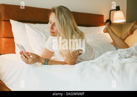 Schöne blonde Frau mit Smartphone, die im Schlafzimmer auf dem Bett liegt Stockfoto