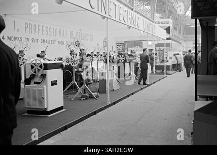 Besucher einer Fachmesse für Film- und Fotohersteller und Zulieferer in Olympia, West Kensington, Hammersmith, London, England, UK in c.1960. Am Stand der Cinetechnic waren professionelle 16-mm-Kinoprojektoren zu sehen. Dahinter befindet sich ein Stand von View-Master, einem Unternehmen, das sich auf stereoskopische Betrachter für kleine Farbfotos (auf Plattenrollen aus Karton) spezialisiert hat und heute noch beliebt ist (jetzt von Mattel hergestellt). Das ist von einem alten Schwarzweiß-negativ eines Amateurfotografen – ein Vintage-Foto aus den 1950er/60er Jahren. Stockfoto