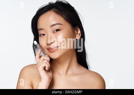 Die junge asiatische Frau hält Hautpflegeprodukte nahe an ihrem Gesicht und lächelt strahlend und selbstbewusst. Stockfoto