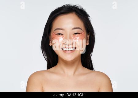 Eine helle Atmosphäre zeigt eine fröhliche junge Frau, die während ihrer Hautpflege Augenflecken aufträgt. Stockfoto