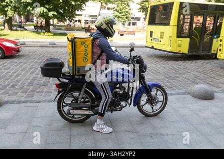 LVIV, UKRAINE - 20. Juni 2021: Ein Fahrer mit Lebensmittellieferanten wird auf einem blauen Motorrad mit einem gelben Rucksack auf der Straße gesehen, der sich auf die Lieferung von Lebensmitteln vorbereitet. Die Stadt ist lebhaft mit Fußgängern und Bussen. Stockfoto
