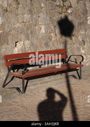 Das Bild zeigt eine Holzbank mit Armlehnen und Beinen aus Metall, die auf einer Kopfsteinpflasteroberfläche platziert sind. Er wird an einer Steinmauer positioniert. Am Boden, Stockfoto