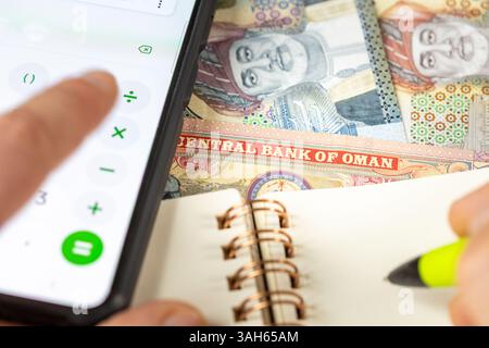 Oman Rial zählt auf Smartphone-Rechner, OMR-Geld, Notizen im Notizbuch. Finanzielle Abrechnung, Haushaltsbudget, Steuern, omanischer Währungsrat Stockfoto