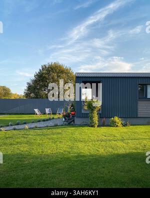 Hinterhof in Sonnenstrahlen am Nachmittag: Drei Liegestühle auf einem Rasen, Veranda mit Sommerhaus und Fußweg. Mehrere Bäume hinter dem grauen Zaun. Stockfoto