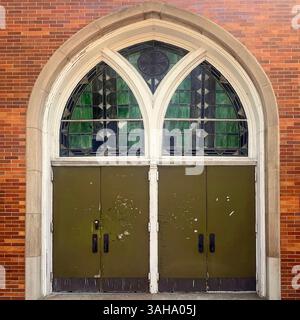 Alte grüne Kirchtüren unter einem großen gotischen Bogen mit Buntglasfenstern in geometrischen Mustern, in einer roten Backsteinfassade in Chicago Stockfoto