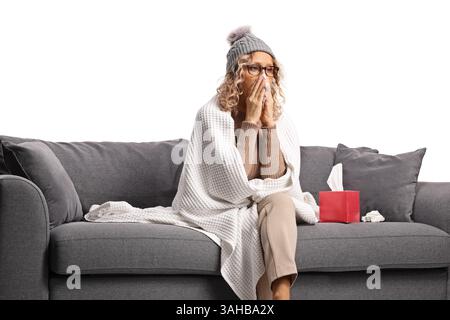 Frau, die in eine Decke gewickelt ist und sich krank fühlt, Nase bläst und auf einem Sofa sitzt, isoliert auf weißem Hintergrund Stockfoto