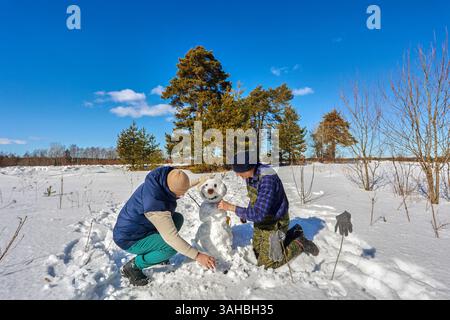 Das Paar in den 50er Jahren arbeitet eng an den letzten Schliffen des Schneemanns, der in der Nähe einer Gruppe von Kiefern Arme und Gesichter hinzufügt, in friedlicher, verschneite Lichtung unter blauem Himmel. Stockfoto