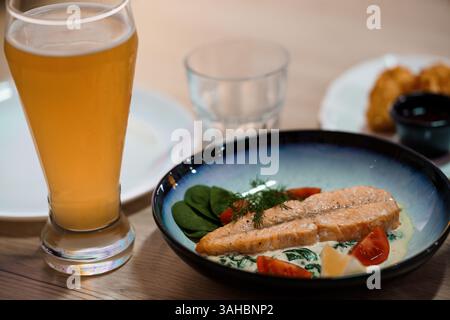 Nahaufnahme eines Glases Bier mit Lachs mit Spinat, Tomaten und Sauce auf einem dunkelblauen Teller in einem Restaurant. Stockfoto