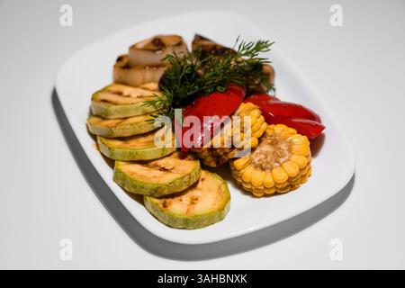 Gegrilltes Gemüse auf einem weißen Teller, einschließlich Zucchini, Zwiebeln, rotem Pfeffer und Mais mit frischer Dill-Beilage Stockfoto