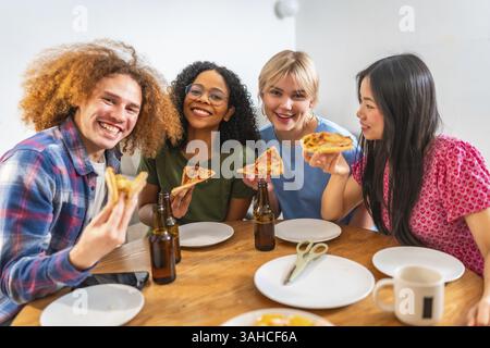 Vier junge, fröhliche Freunde mit unterschiedlichem Hintergrund genießen Pizza und Bier zu Hause, feiern Freundschaft und gute Zeiten Stockfoto