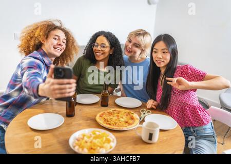 Vier verschiedene Freunde machen ein Selfie mit einem Smartphone, während sie zu Hause ein Mittagessen mit Pizza und Bier genießen Stockfoto