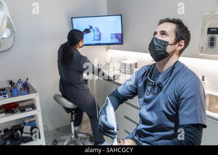 Zahnarzt, der mit einer UV-Lampe eine Zahnaufhellung durchführt, wobei eine Zahnarzthelferin im Hintergrund eine Röntgenaufnahme analysiert Stockfoto