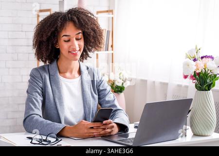 Tolle Bewertungen von Kunden bis hin zu Online-Modegeschäften für kleine Unternehmen. Lächelnde afroamerikanerin liest im Smartphone und sitzt mit am Tisch Stockfoto