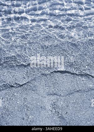 Wunderschöne kleine Meereswellen mit sauberem, transparentem Wasser am Sandstrand der Küste an sonnigen Tagen. Sauberes Wasser, Sonnenblendung und Schatten auf Sandboden in Nahaufnahme. Natürlicher abstrakter Hintergrund. Stockfoto