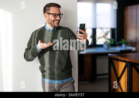 Selbstbewusster, lächelnder Geschäftsmann mit Smartphone im Heimbüro. Überprüfen neuer E-Mails, Videoanrufe Stockfoto